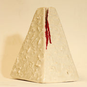 Offwhite ceramic sculpture of a pyramid shaped vase, with a wide base and a slightly narrow top. The vase is bumpy in texture and has a subtle cat face on the top. It has streaks of red running down the side of it.