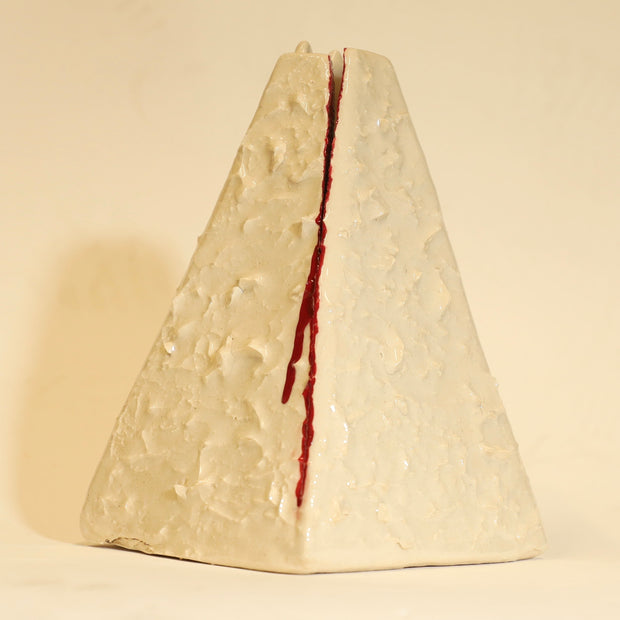 Offwhite ceramic sculpture of a pyramid shaped vase, with a wide base and a slightly narrow top. The vase is bumpy in texture and has a subtle cat face on the top. It has streaks of red running down the side of it.