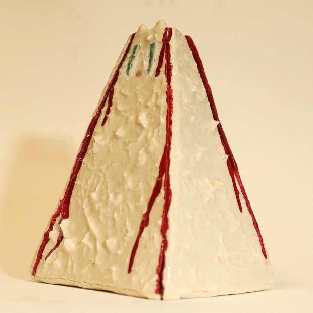 Offwhite ceramic sculpture of a pyramid shaped vase, with a wide base and a slightly narrow top. The vase is bumpy in texture and has a subtle cat face on the top. It has streaks of red running down the side of it.