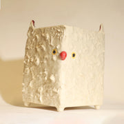 Box shaped ceramic vessel, off white in coloring with a set of small yellow and black eyes and a round red nose. It has 2 pointed ears and small feet.