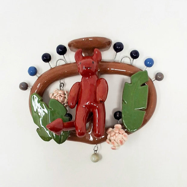 Ceramic sculpture of a tree of life with a red devil as the center and leaves and flowers hanging off it. Several rounded balls stick out of the vessel with wire.