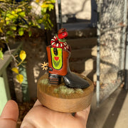 Sculpture of a red snake with black and white stripes, coiled up inside of a brown and black cowboy boot with a spur. Cowboy boot has a design element of a pink tulip with yellow stars on green background on each side of the boot. Snake wears a small cowboy hat and bandana. 