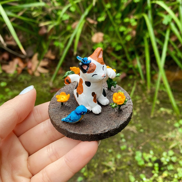 Sculpture of a calico cat sitting with one paw raised and a closed eye, smiling expression. A blue butterfly sits atop its head. The cat holds an orange flower in its tail and sits on a brown dirt circular base with orange flowers and a blue and orange bird.