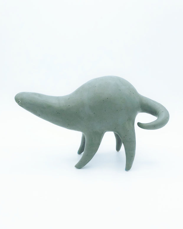 Ceramic sculpture of a rounded body character, a muted teal in color with  simple line smile and small eyes. It has a long neck and a long skinny tail.