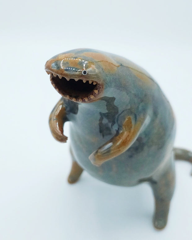 Glazed ceramic sculpture of a large rounded body monster, standing on hind legs with many small teeth and an open mouth. Colors are blue with brown accent coloring.