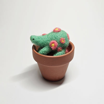 Needle felted wool sculpture of a green rounded body quadruped creature with an cute smile and small black dot eyes. It is positioned in a clay pot and has pink flowers attached to its body.