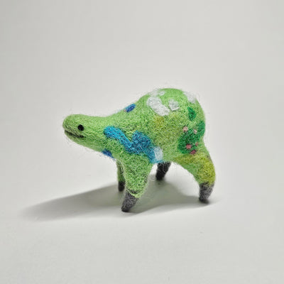 Needle felted wool sculpture of a green rounded body quadruped creature with a cute smile and small black dot eyes. Its body has patterns of clouds, water, and flowers like its a miniature world.