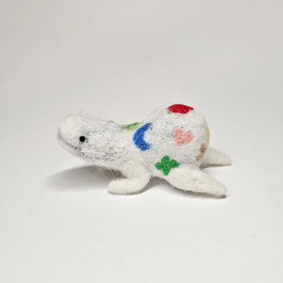 Needle felted wool sculpture of a white rounded body quadruped creature with a cute smile and small black eyes. It lays on its stomach and has colorful icons on its body: heart, crescent moon, clover, red balloon, etc.