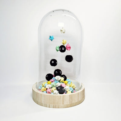 Glass cloche with a wooden bottom containing many small sculptures of black soot sprites made out of wool. They hang from the top of the glass and are surrounded by paper stars.