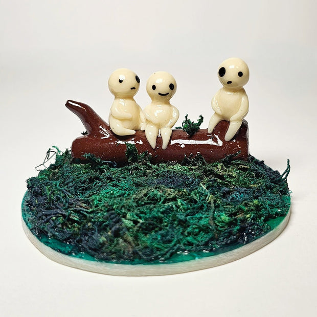 Sculpture of 3 small white ghostly spirit characters (Kodama from Princess Mononoke) sitting atop of a log, which is positioned on a circular mossy base.