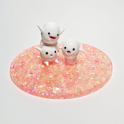 Sculpture of 3 small white characters with round bodies, tiny arms and legs, and cute cartoon expressions. They stand atop a pink glittery circular base.