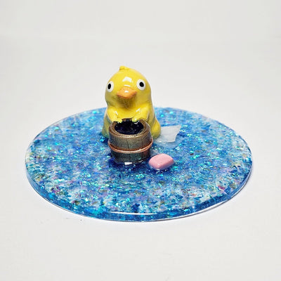 Sculpture of a yellow duck standing over a small wooden bucket  with a wash cloth behind it and a bar of pink soap to the side. The sculpture is a part of a glittery blue circular resin base.