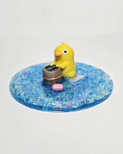 Sculpture of a yellow duck standing over a small wooden bucket  with a wash cloth behind it and a bar of pink soap to the side. The sculpture is a part of a glittery blue circular resin base.