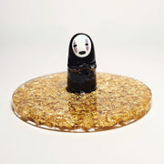 Sculpture of No Face, with his head slightly tilted and standing atop of a circular gold sparkly base made out of resin. He holds out his hands.