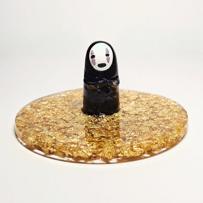 Sculpture of No Face, with his head slightly tilted and standing atop of a circular gold sparkly base made out of resin. He holds out his hands.