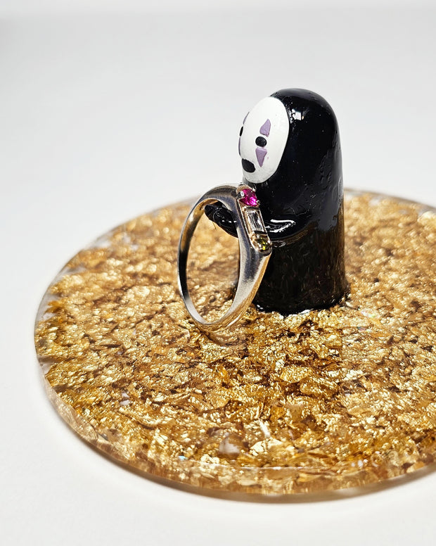 Sculpture of No Face, with his head slightly tilted and standing atop of a circular gold sparkly base made out of resin. He holds out his hands and a ring is placed on them.