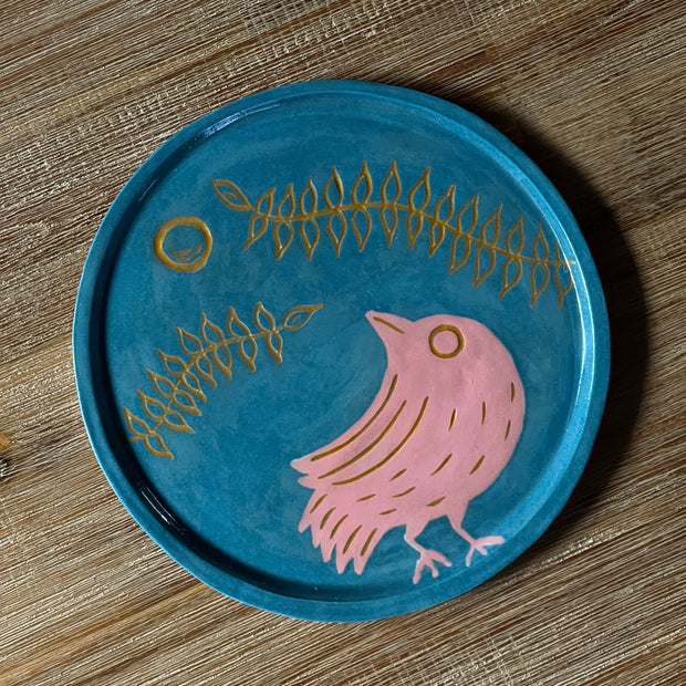 Blue ceramic plate with an inlayed illustration of a pink bird looking up and off to the side. Over it is a circular moon and 2 long vines with several leaves.