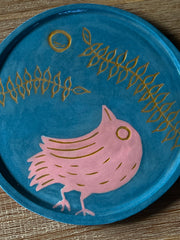 Blue ceramic plate with an inlayed illustration of a pink bird looking up and off to the side. Over it is a circular moon and 2 long vines with several leaves.