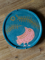 Blue ceramic plate with an inlayed illustration of a pink bird looking up and off to the side. Over it is a circular moon and 2 long vines with several leaves.