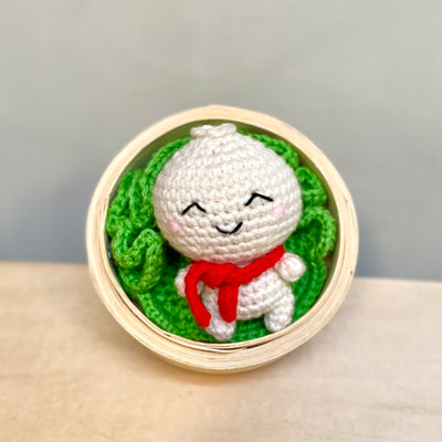 Crochet sculpture of a bao bun with a cute, smiling face and a tiny body. It wears a red scarf and sits in a crocheted bed of lettuce inside of a real steamer basket.