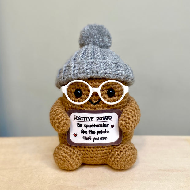 Crochet sculpture of a brown potato character with a set of black beaded eyes and a small smile. It wears white glasses, a gray beanie and holds a sign that reads "Positive Potato"