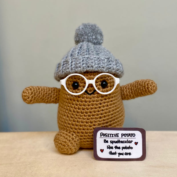 Crochet sculpture of a brown potato character with a set of black beaded eyes and a small smile. It wears white glasses, a gray beanie and extends out its arms. Nearby is a sign that reads "Positive Potato"