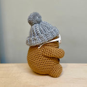 Side view of crochet potato character with short arms and legs, a gray pompom beanie and white glasses. 