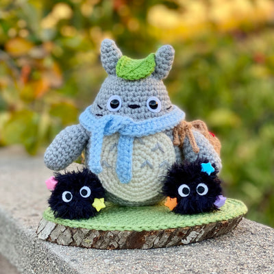 Crochet knitting sculpture of Totoro, wearing a blue scarf and a green leaf atop his head next to 2 soot sprites with stars attached. They are atop of a wood slice base with a green crochet rug atop.