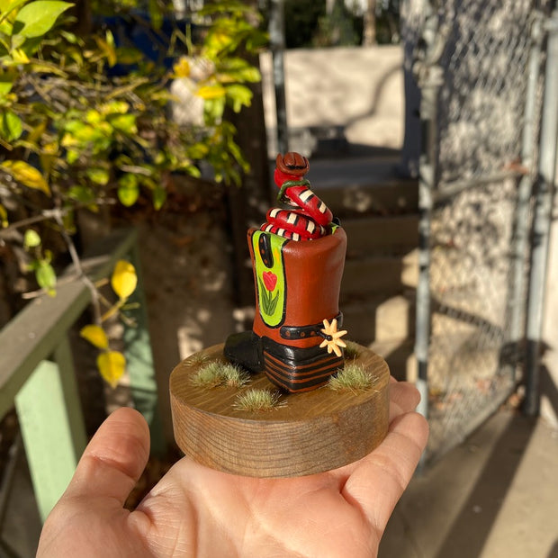 Sculpture of a red snake with black and white stripes, coiled up inside of a brown and black cowboy boot with a spur. Cowboy boot has a design element of a pink tulip with yellow stars on green background on each side of the boot. Snake wears a small cowboy hat and bandana. 