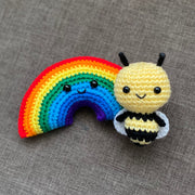 2 crocheted plushes: one of a rainbow and the other of a bee with a large cartoon like head. They both have black bead eyes and small smiles.