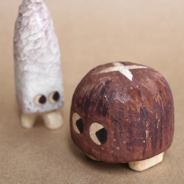 Group of 2 small mushrooms whittled out of wood. One is tall and white, the other is short and brown like a shiitake. Both have cartoon eyes and very short legs.