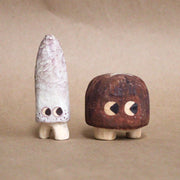 Group of 2 small mushrooms whittled out of wood. One is tall and white, the other is short and brown like a shiitake. Both have cartoon eyes and very short legs.