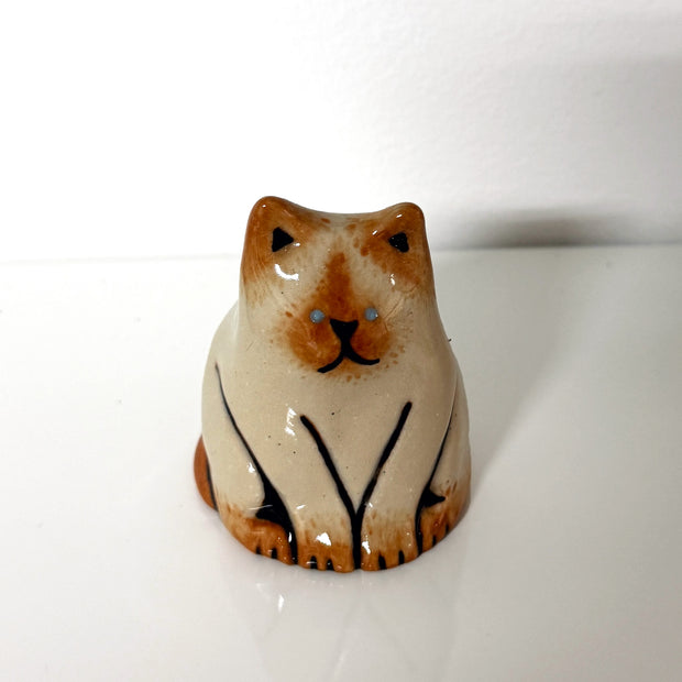 Small ceramic sculpture of a tan cat with warm brown colored ears, paws, tail and snout. It sits and looks forward with blue eyes and a slightly smiling expression.