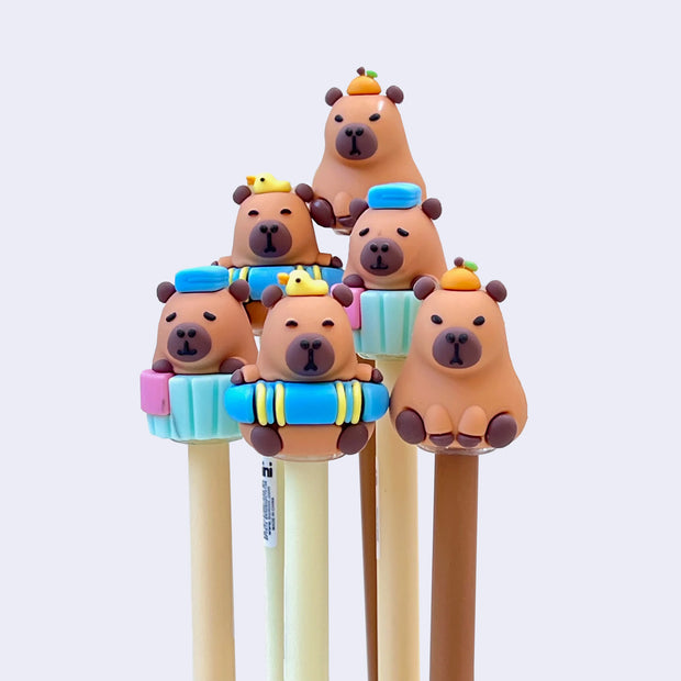 Group of gel pens with rubber capybaras as pen toppers. Capybaras are either: soaking in a bath with a towel atop their head, in a rubber floaty, or sitting with a tangerine atop its head.