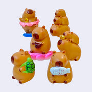 6 options of capybara figurines, in different poses. Options are: dancing w/ tutu, stacked, playing wood instrument, stacked w/ gold, turtle backpack and holding fish.