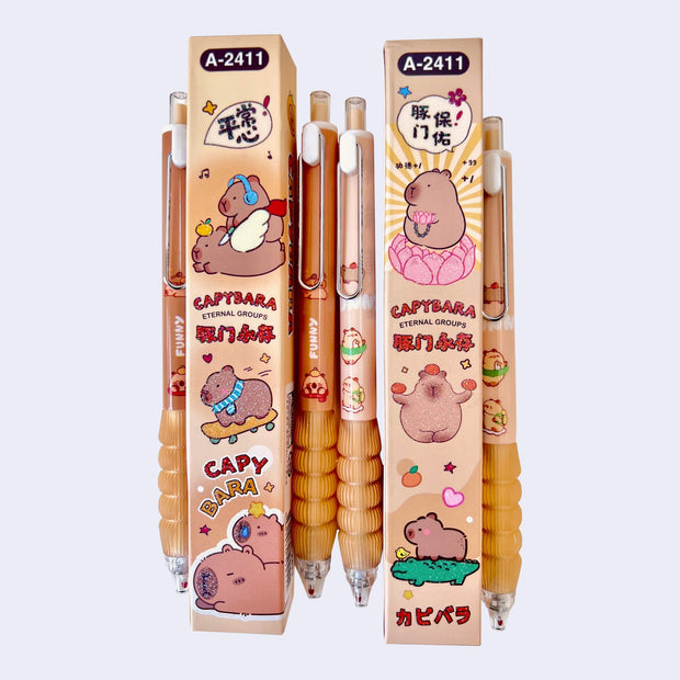 Collection of several gel pens with brown bodies, a rounded metal clip and a squishy barreled surface for placing one's hand. Small illustrations of cartoon capybaras are on the pens and product packaging.