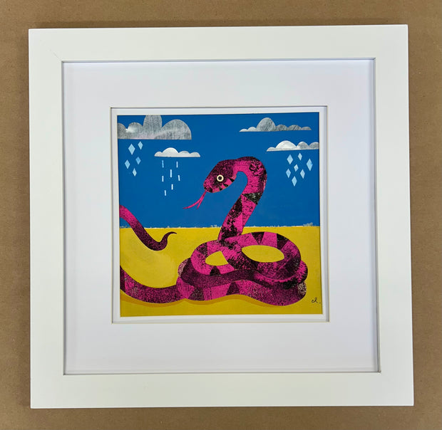 Paper collage art of a pink snake with black dotting and striping, with much of its body coiled up. It has its tongue out and sits in a yellow land with blue sky and raining clouds. Piece is in white frame.