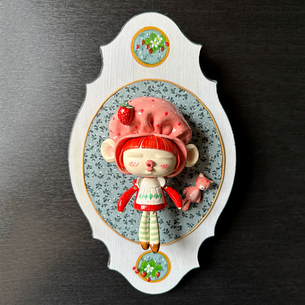 Ceramic sculpture of a doll with a cute, large closed eye head. It is dressed like Strawberry Shortcake and mounted to a wood plaque with a small bear.