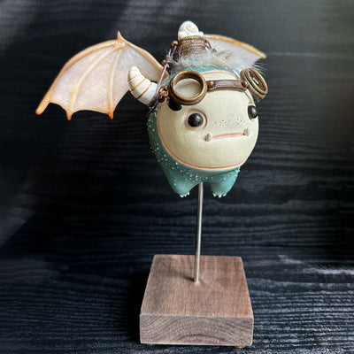 Sculpture of a large headed teal monster with leather goggles atop its head and large bat wings. Its mounted via a metal rod to a square block and has a tiny character in a basket atop its head, riding it.