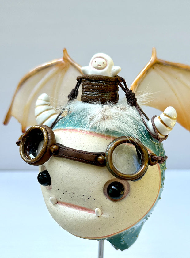 Sculpture of a large headed teal monster with leather goggles atop its head and large bat wings. Its mounted via a metal rod to a square block and has a tiny character in a basket atop its head, riding it.