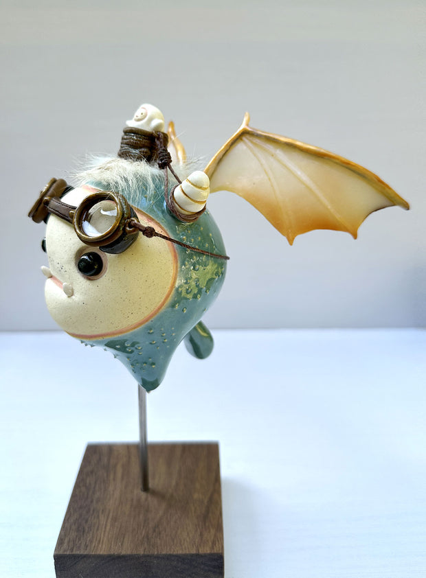 Sculpture of a large headed teal monster with leather goggles atop its head and large bat wings. Its mounted via a metal rod to a square block and has a tiny character in a basket atop its head, riding it.