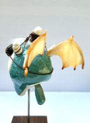 Sculpture of a large headed teal monster with leather goggles atop its head and large bat wings. Its mounted via a metal rod to a square block and has a tiny character in a basket atop its head, riding it.