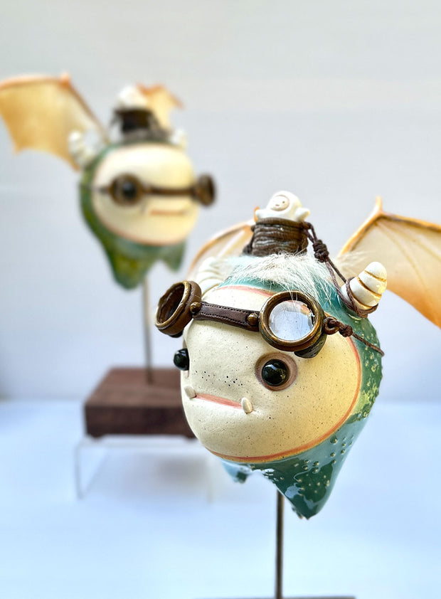 Sculpture of a large headed teal monster with leather goggles atop its head and large bat wings. Its mounted via a metal rod to a square block and has a tiny character in a basket atop its head, riding it.