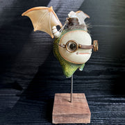 Sculpture of a large headed green monster with leather goggles atop its head and large bat wings. Its mounted via a metal rod to a square block and has a tiny character in a basket atop its head, riding it.