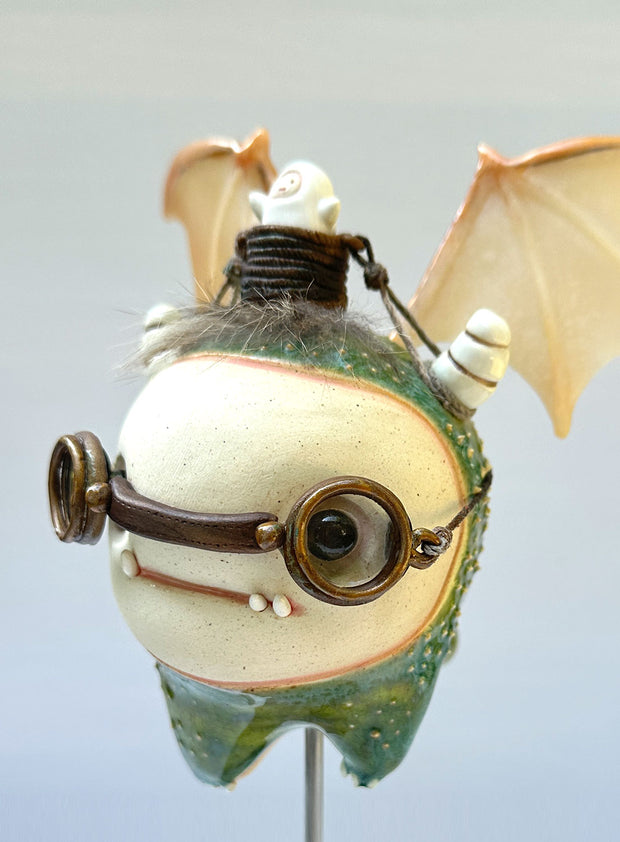 Sculpture of a large headed green monster with leather goggles atop its head and large bat wings. Its mounted via a metal rod to a square block and has a tiny character in a basket atop its head, riding it.