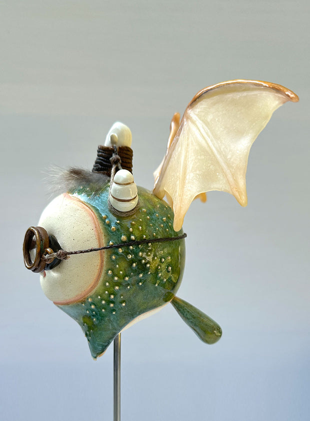 Sculpture of a large headed green monster with leather goggles atop its head and large bat wings. Its mounted via a metal rod to a square block and has a tiny character in a basket atop its head, riding it.