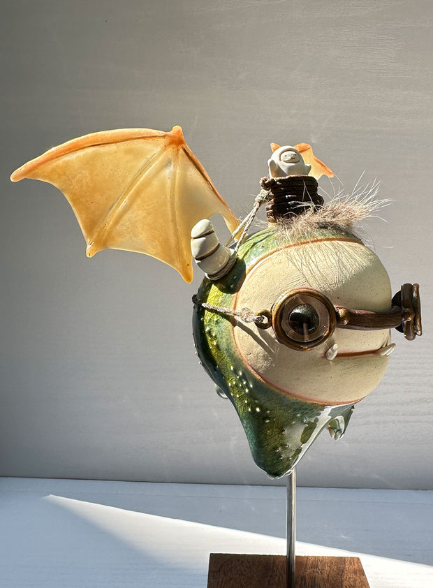 Sculpture of a large headed green monster with leather goggles atop its head and large bat wings. Its mounted via a metal rod to a square block and has a tiny character in a basket atop its head, riding it.