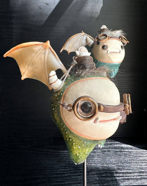 Sculpture of a large headed green monster with leather goggles atop its head and large bat wings. Its mounted via a metal rod to a square block and has a tiny character in a basket atop its head, riding it.