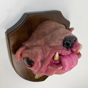 Sculpture of a pink dog's head with a large underbite and a shiny nose and large eyes. Its tongue sticks out and it's head is mounted to a wooden plaque, like a commemorative deer head.