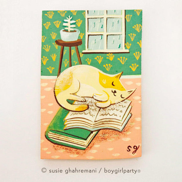 Painting of a white and yellow cartoon cat sleeping on a pile of books in a room with a rainy day window and bright wallpaper and flooring.